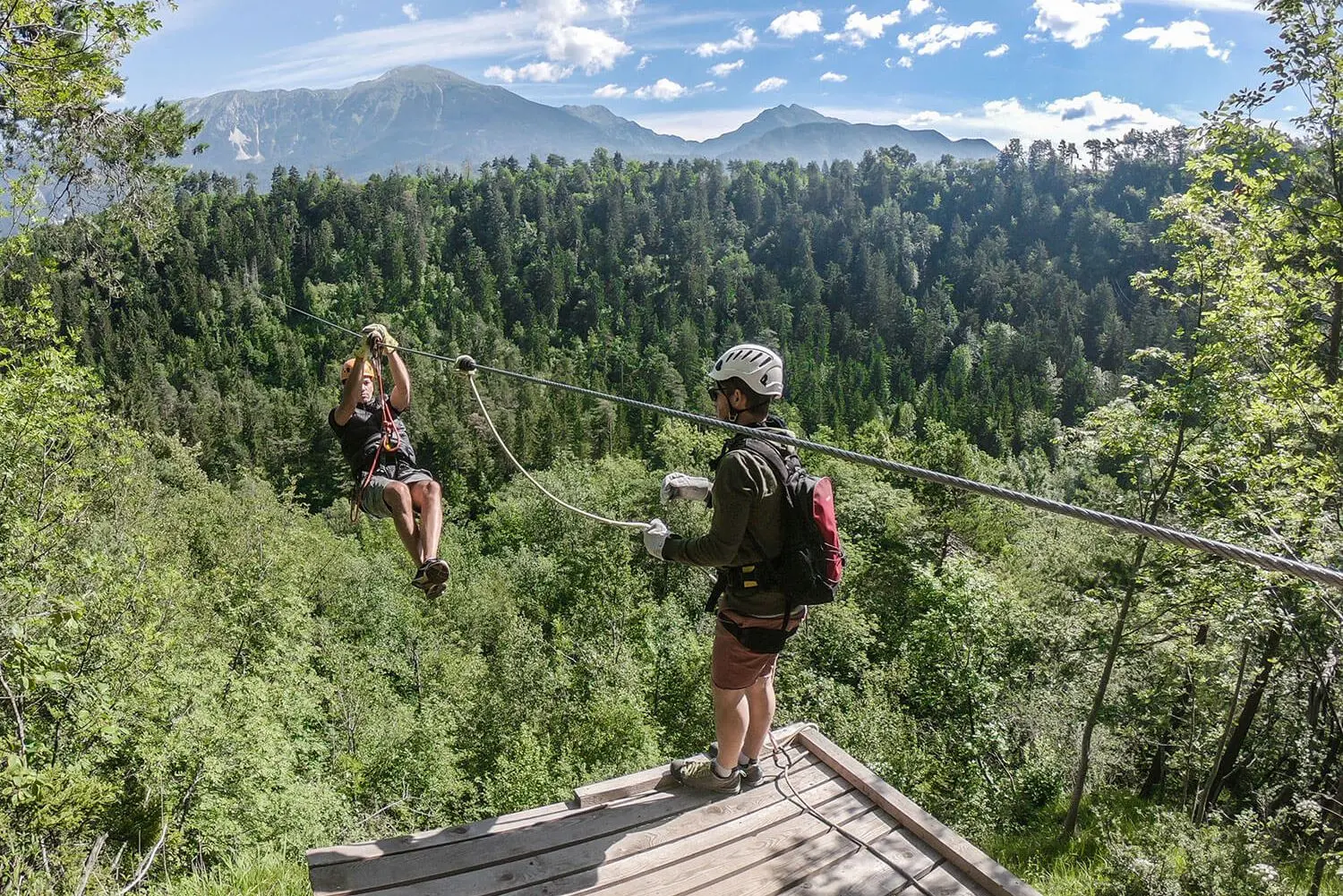 Man zip-lining on a sunny day with scenic forest and mountain background